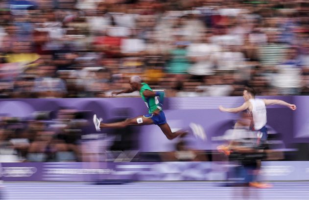 Atletas em prova de atletismo: um salta no ar, o outro corre. Fundo de público borrado por movimento.