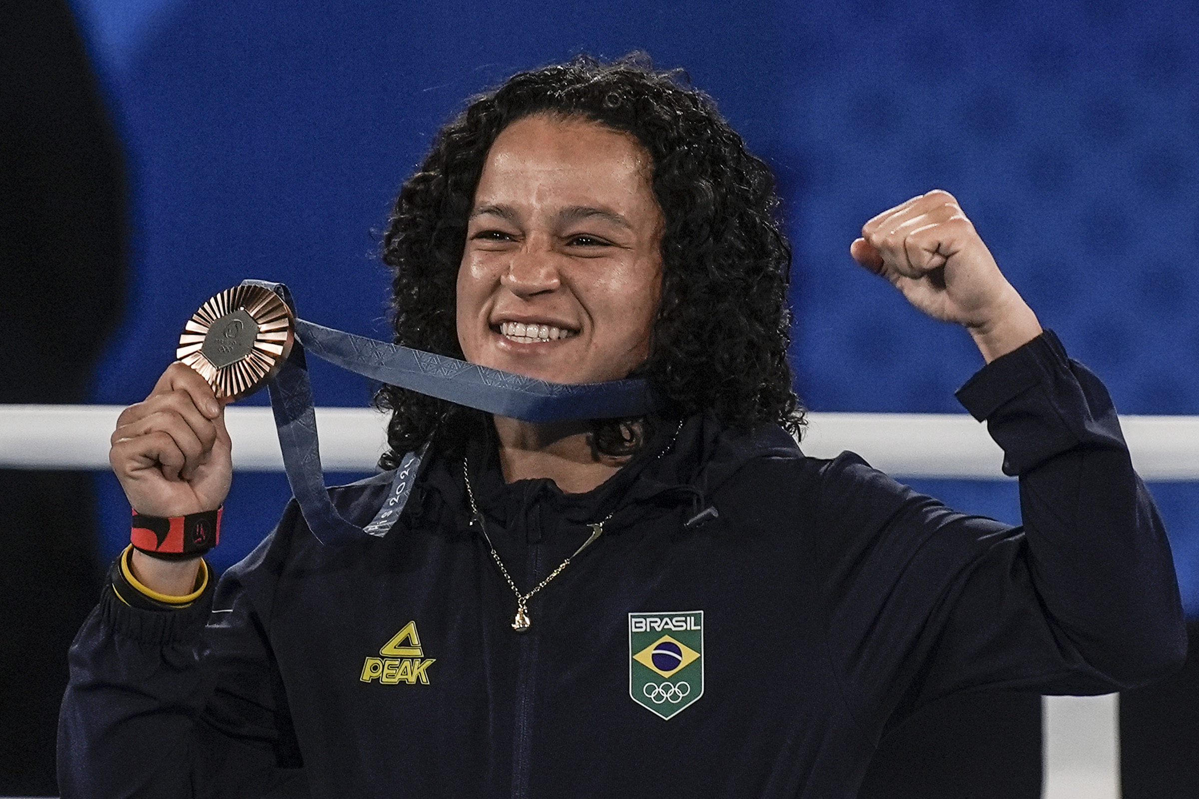 Atleta brasileira sorrindo, exibindo medalha de bronze no pescoço e punho cerrado em celebração.