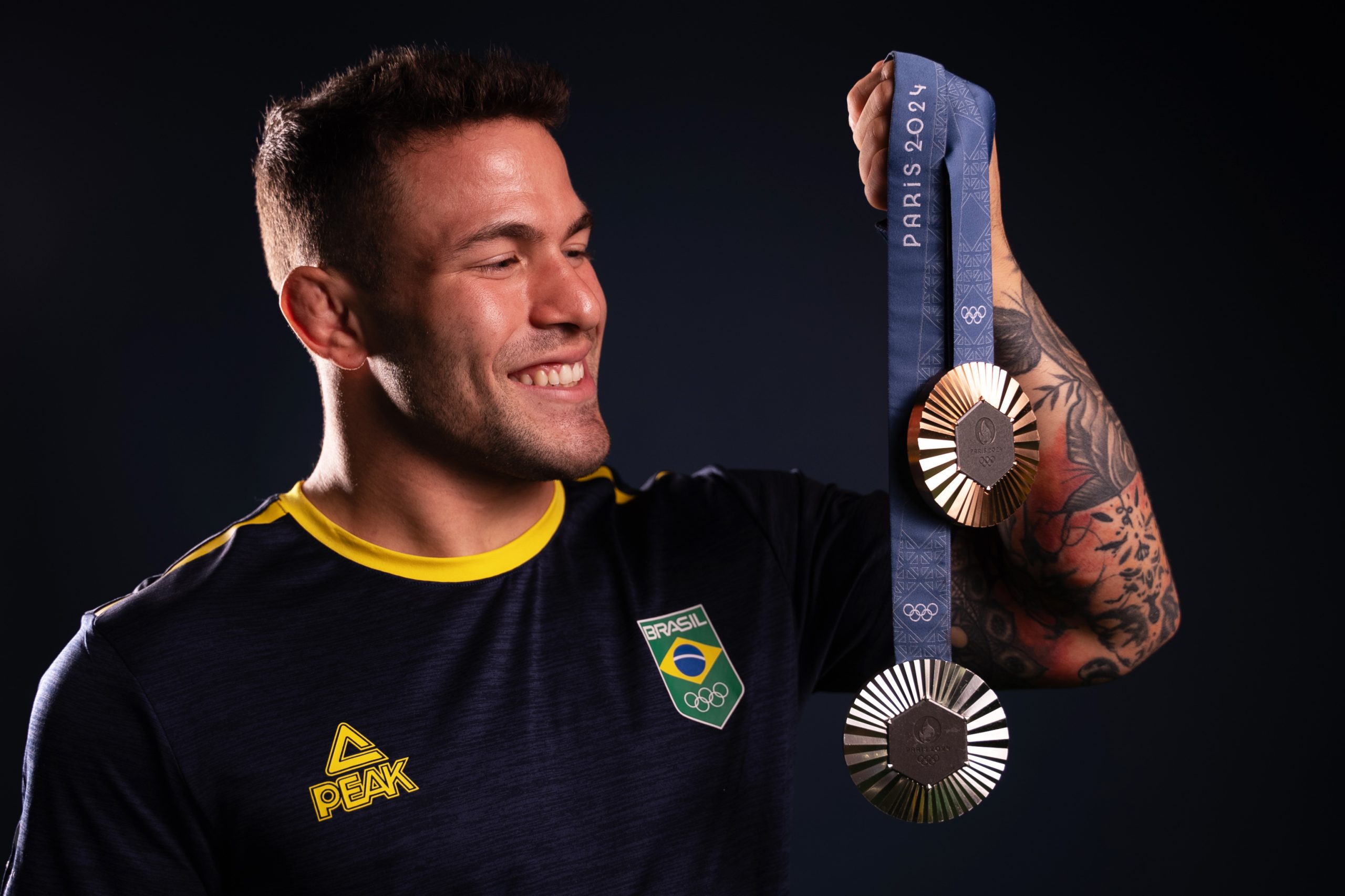 Atleta brasileiro sorrindo e exibindo duas medalhas: uma dourada e uma prateada.