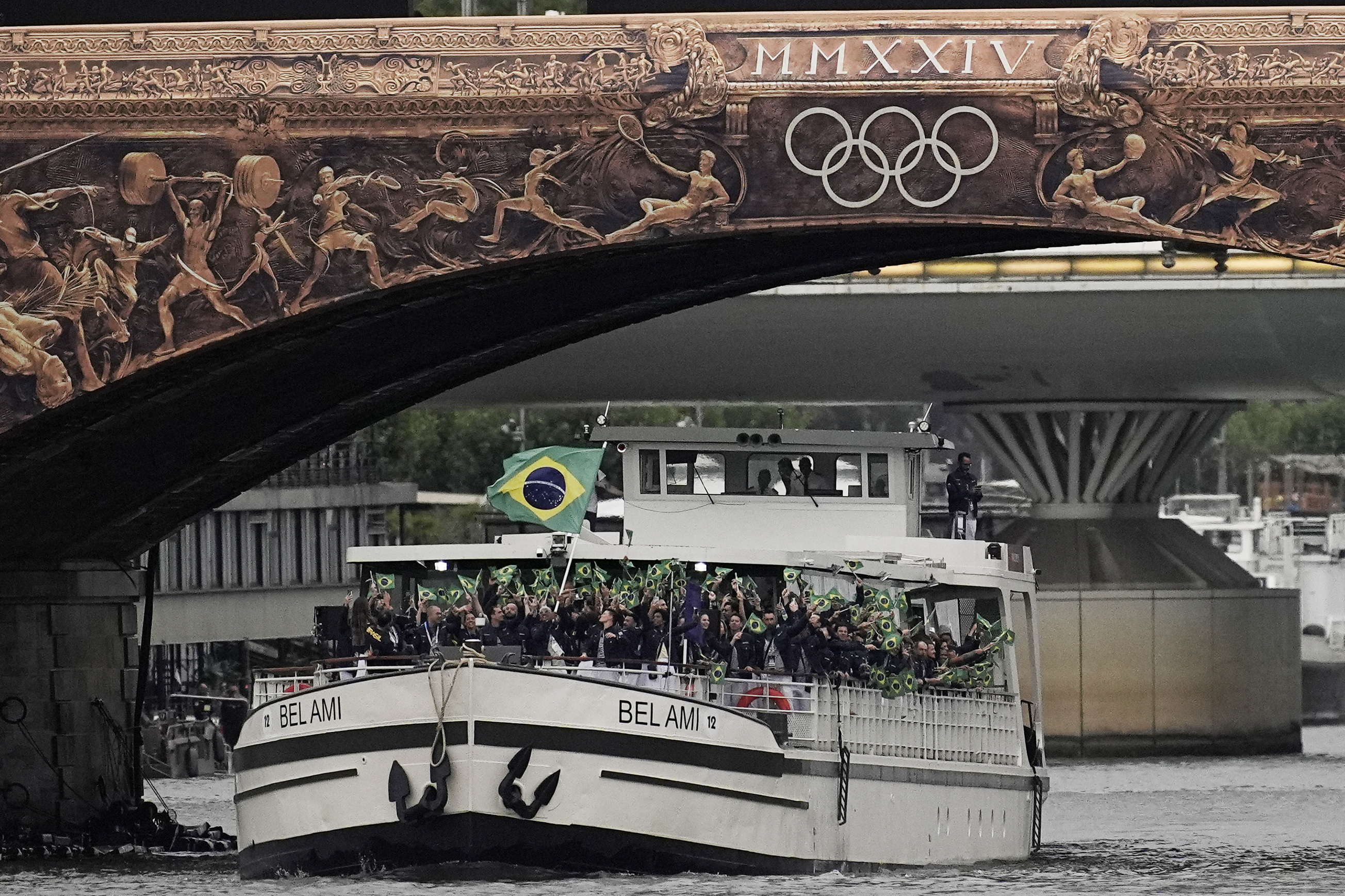 Barco com torcida brasileira acenando bandeiras sob ponte decorada com relevos de atletas e anéis olímpicos.