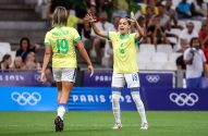 Duas jogadoras de futebol brasileiras no campo, uma de costas (Priscila 19) e outra (18) celebrando. Ao fundo, Paris 2024.