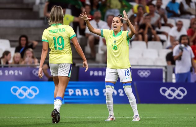 Duas jogadoras de futebol brasileiras no campo, uma de costas (Priscila 19) e outra (18) celebrando. Ao fundo, Paris 2024.