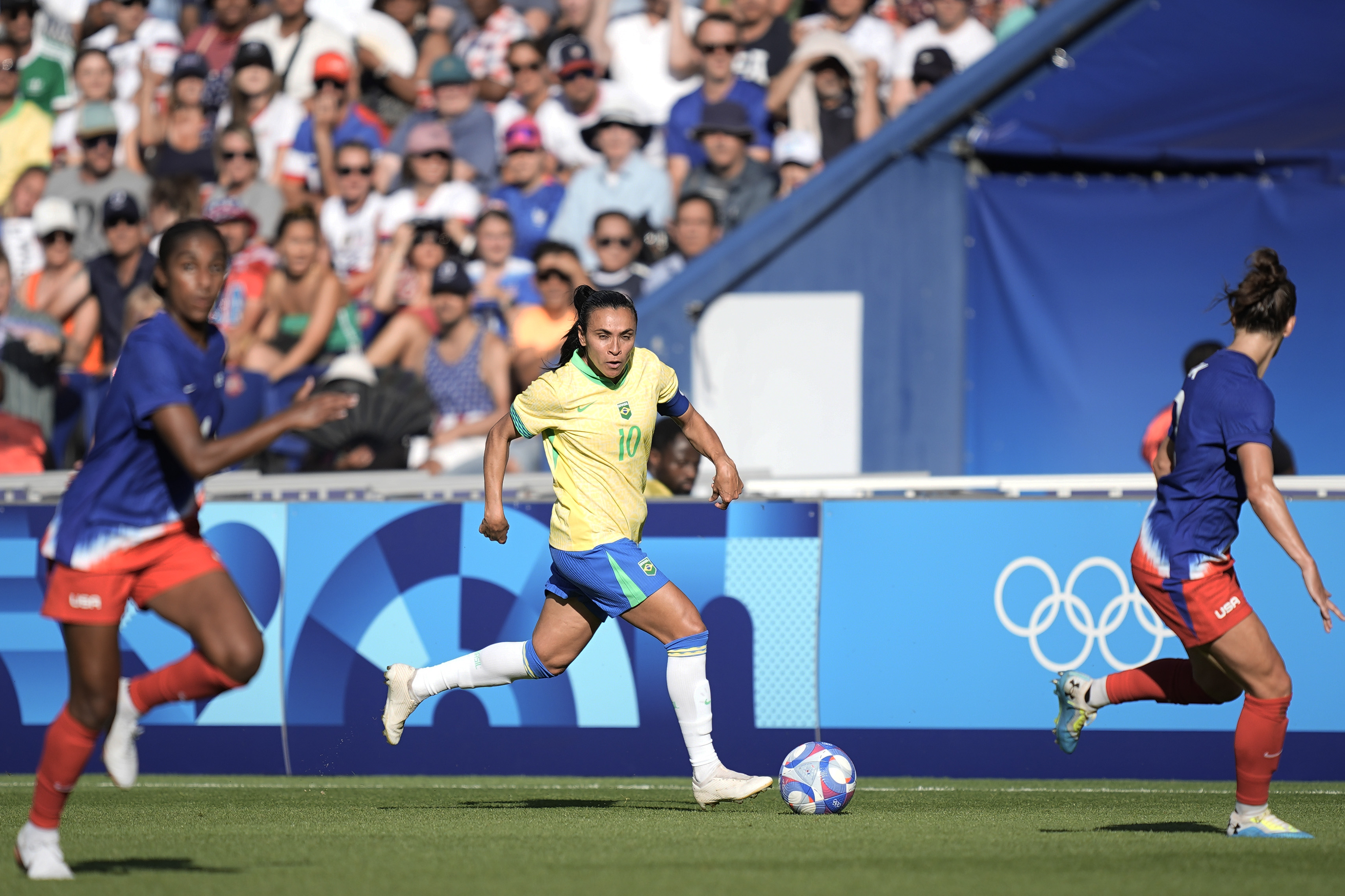 Partida de futebol feminino: Jogadora do Brasil (10) dribla a bola, com adversárias e anéis olímpicos.