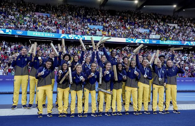 Equipe feminina brasileira celebra com medalhas de prata e estojos no pódio de estádio, com "PARIS 2024".