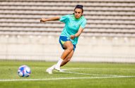Jogadora de futebol brasileira dribla a bola em campo verde, com arquibancadas ao fundo.