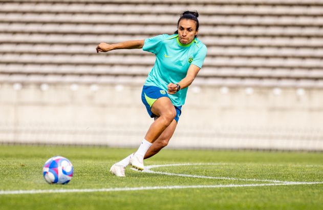 Jogadora de futebol brasileira dribla a bola em campo verde, com arquibancadas ao fundo.