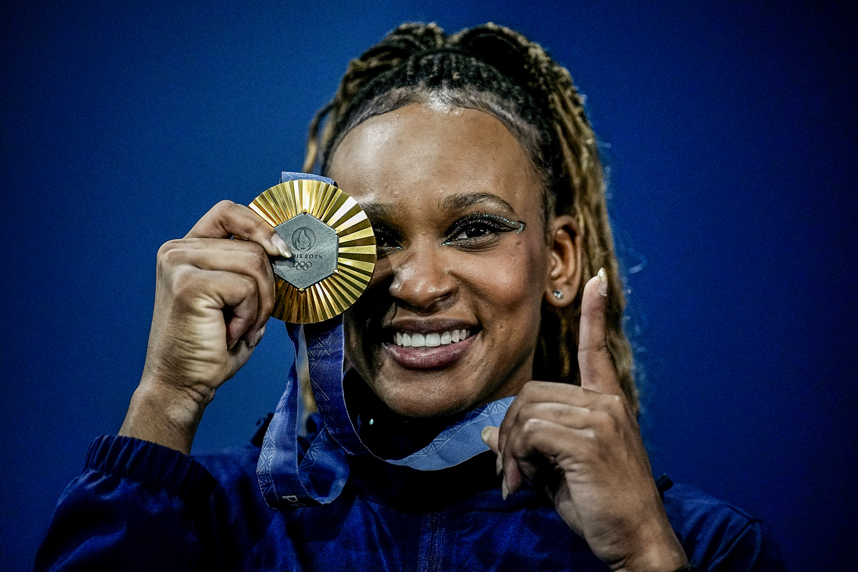 Mulher atleta sorridente com medalha de ouro Paris 2024 sobre o olho e dedo indicador levantado.
