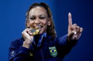 Atleta brasileira morde medalha de ouro, sorrindo e apontando o dedo para cima.