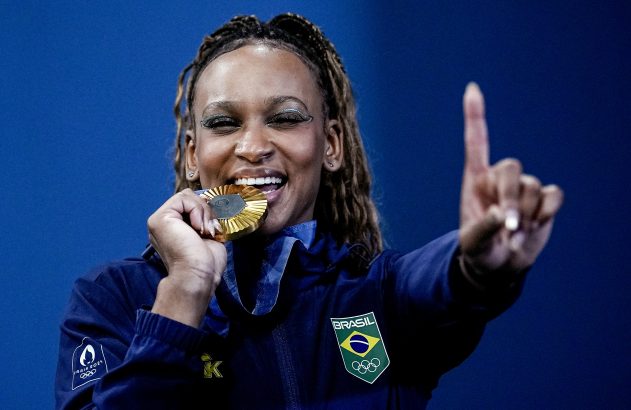 Atleta brasileira morde medalha de ouro, sorrindo e apontando o dedo para cima.