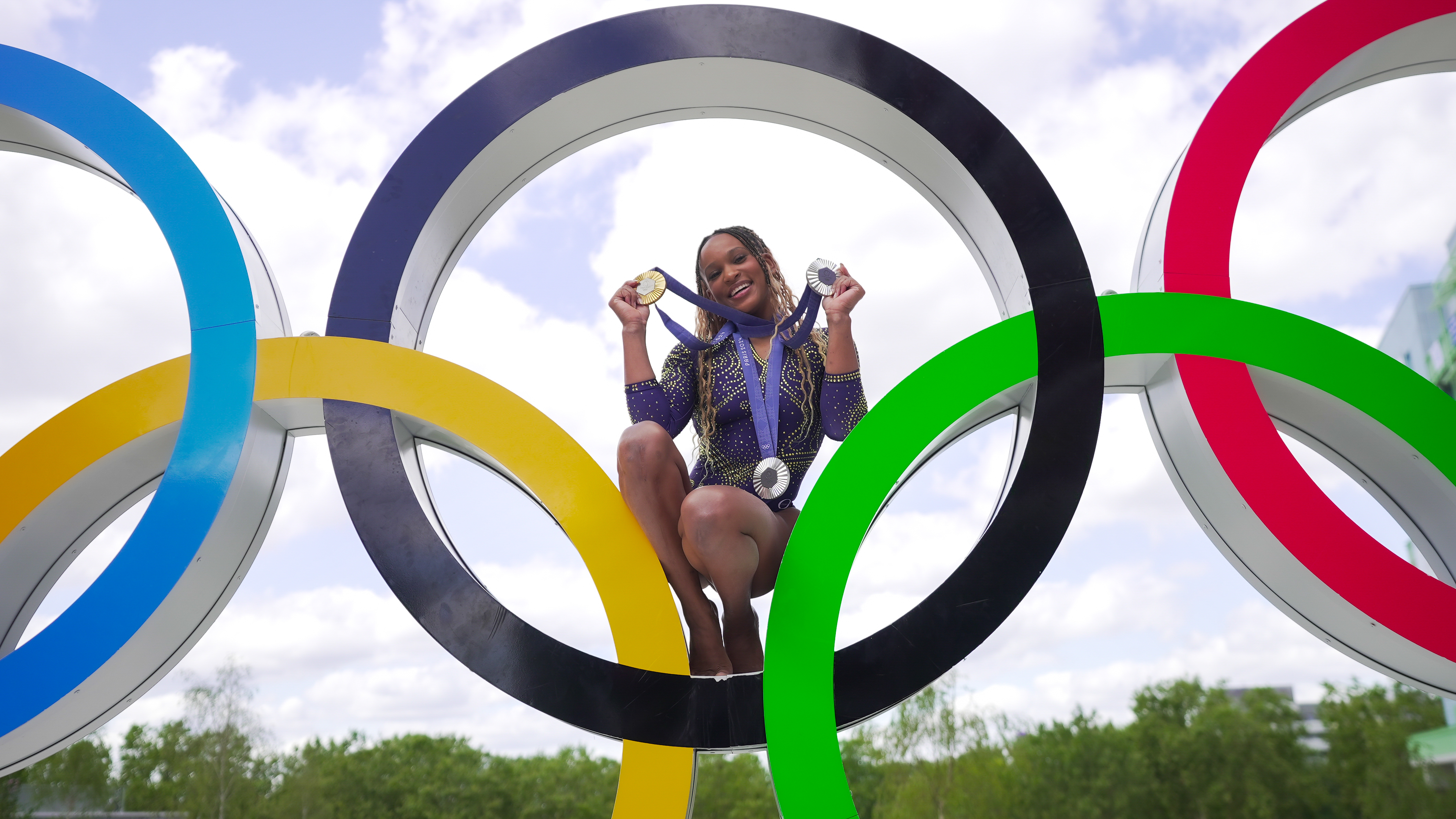 Atleta sorridente posa no anel central dos aros olímpicos, exibindo medalhas.