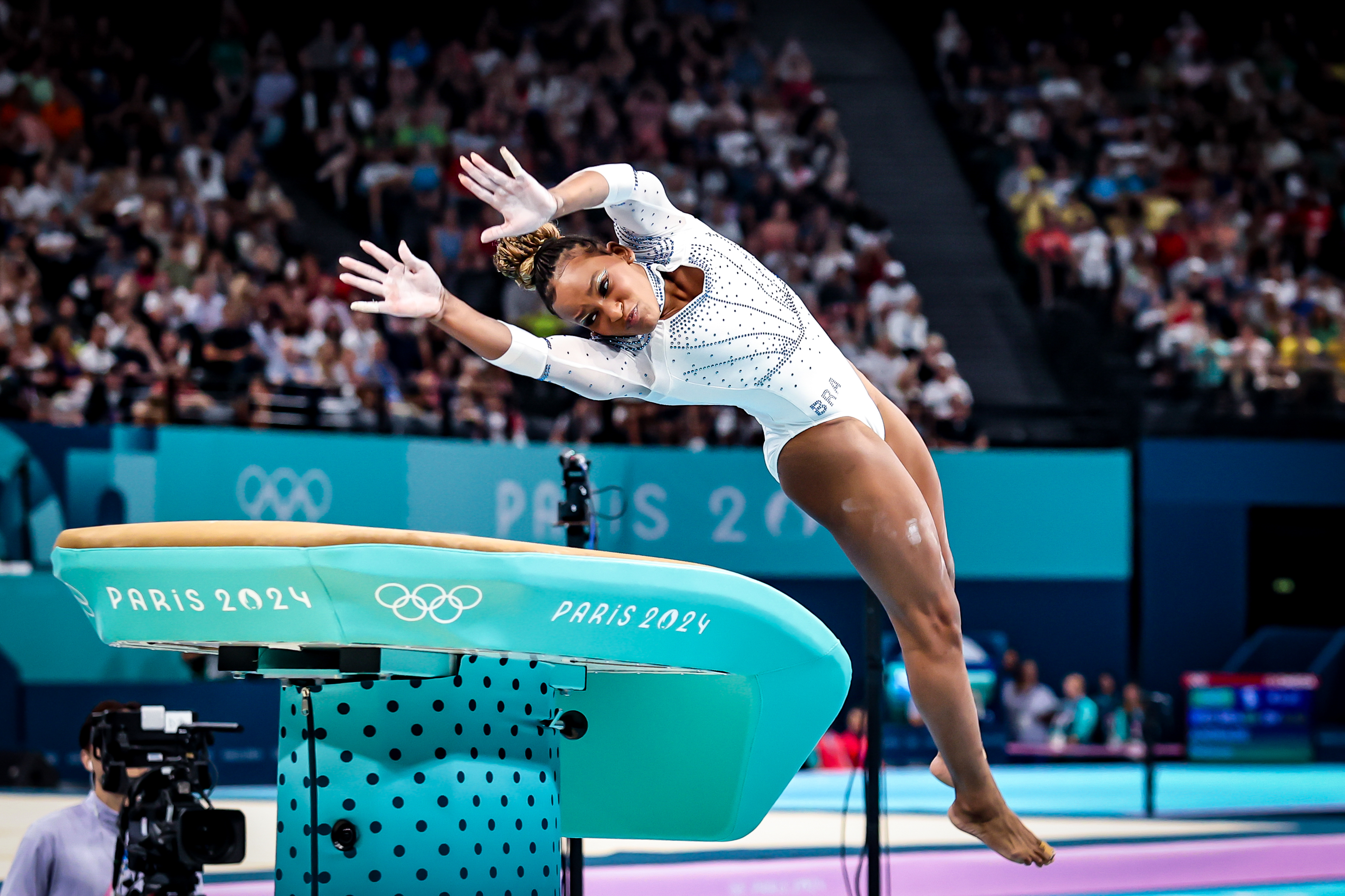 Ginasta, de branco, em pleno salto sobre a mesa com logo Paris 2024. Ginásio olímpico.