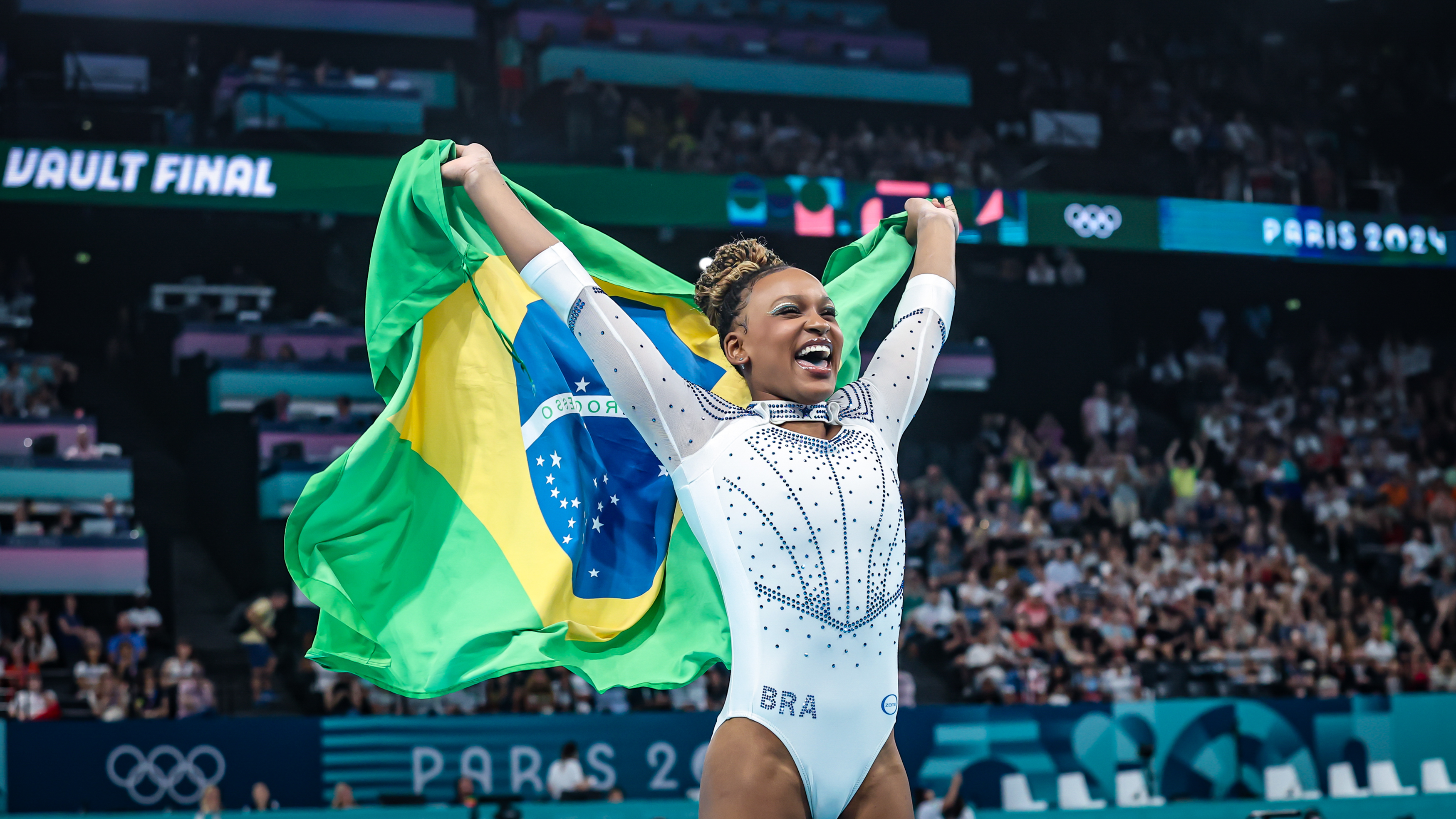 Ginasta brasileira sorridente celebra com bandeira do Brasil erguida em arena.