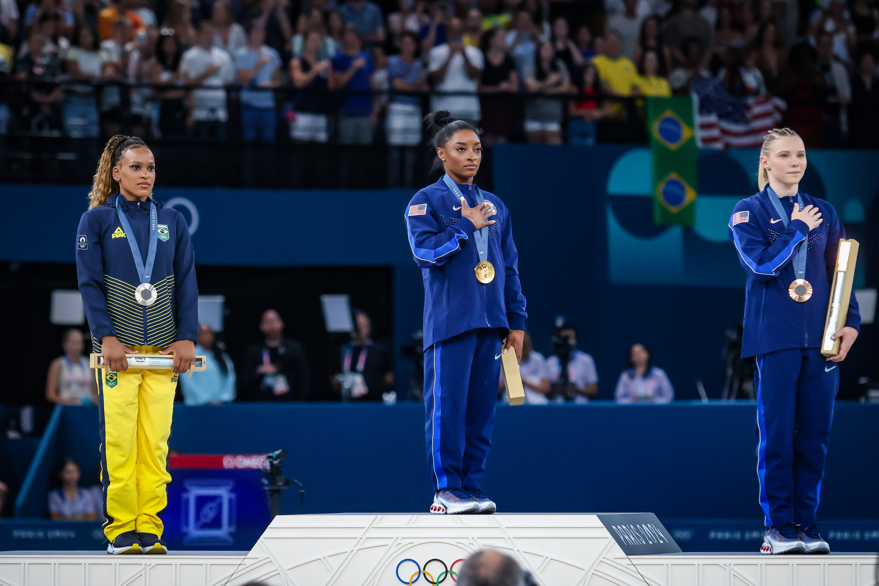 Três ginastas com medalhas (ouro, prata, bronze) no pódio olímpico. Uma delas é Simone Biles. Mão no coração na cerimônia.