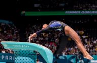 Ginasta salta sobre mesa de salto azul. Arqueada no ar, com "Paris 2024" visível no aparelho.