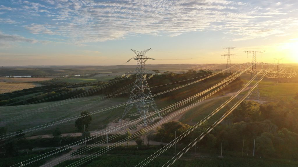 Torres de transmissão e linhas de energia atravessam campos verdes ao pôr do sol.