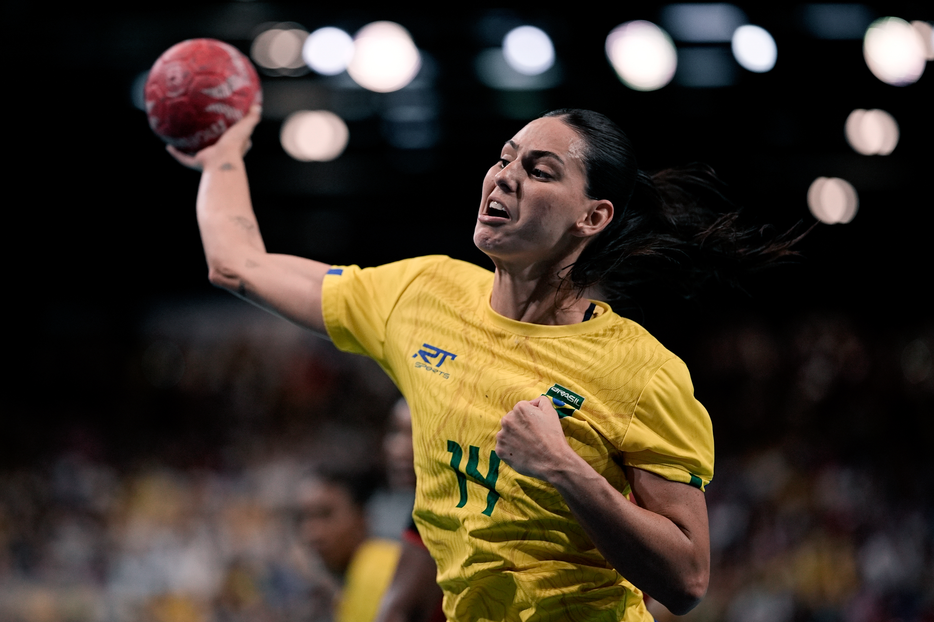 Jogadora de handebol brasileira em camisa amarela e bola vermelha, arremessando com expressão intensa.