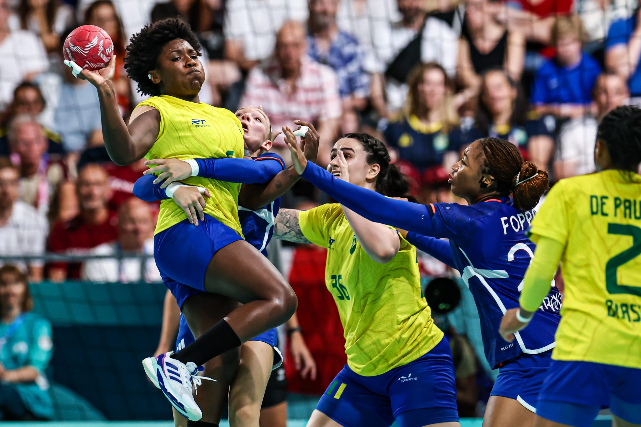 Jogadora de handebol em amarelo salta para lançar bola vermelha. Defensoras em azul tentam bloquear.