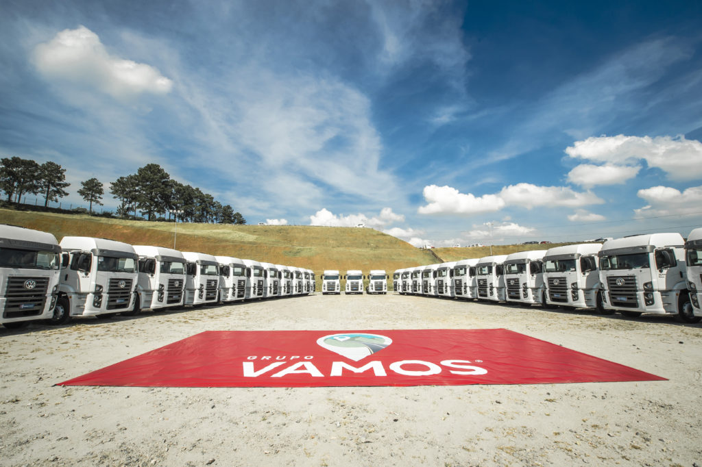 Dezenas de caminhões brancos enfileirados em semicírculo, com banner "Grupo Vamos" em primeiro plano.