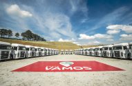 Dezenas de caminhões brancos enfileirados em semicírculo, com banner "Grupo Vamos" em primeiro plano.