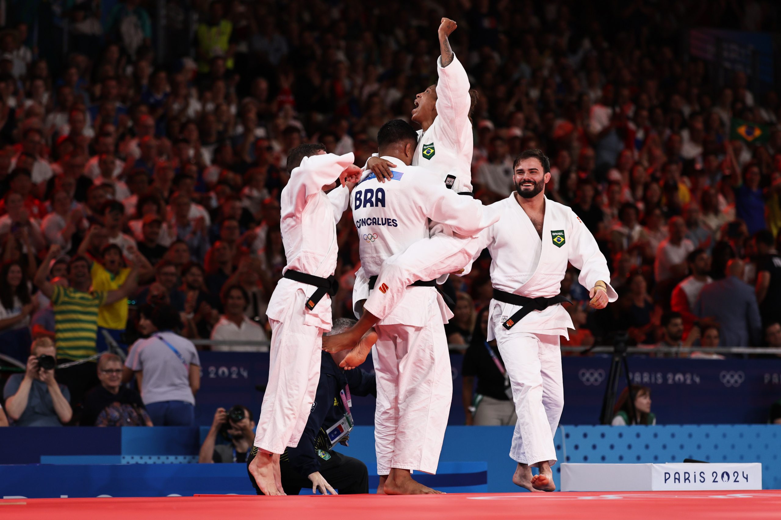 Judocas brasileiros celebram vitória, um é erguido. Público torce. Paris 2024.