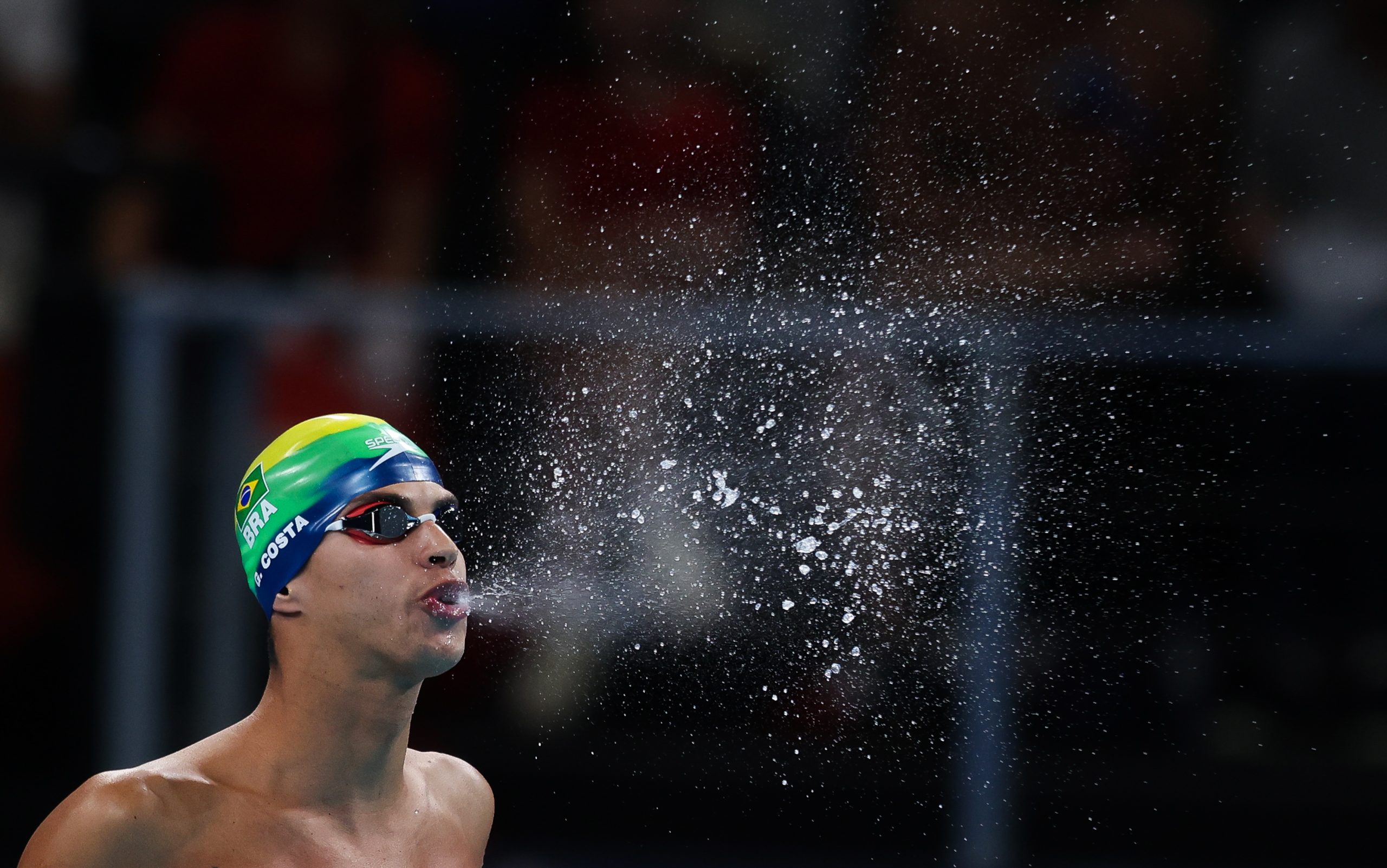 Nadador brasileiro com touca verde e amarela sopra um grande jato de água da boca. Gotas voam no ar contra o fundo escuro.