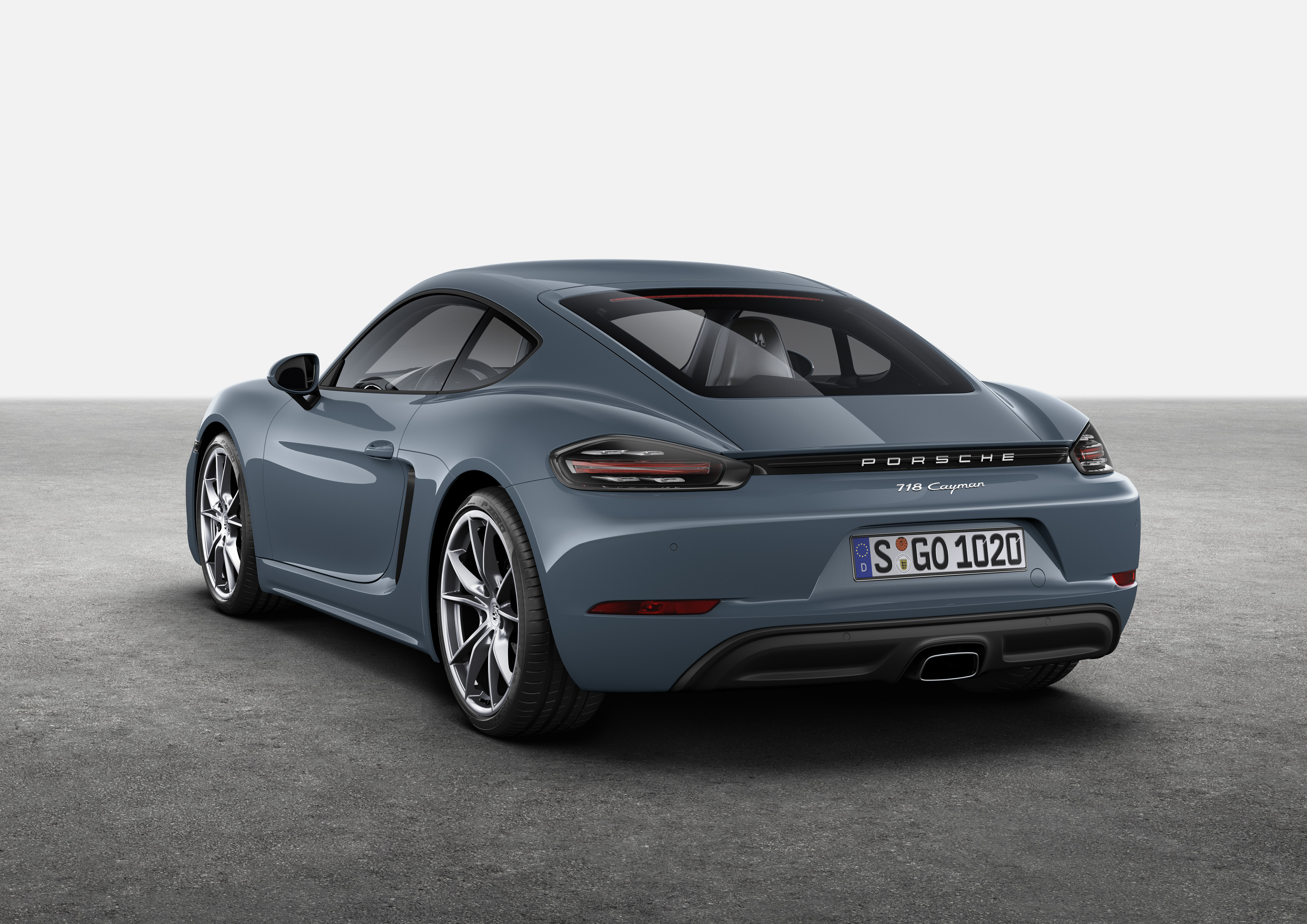 Porsche 718 Cayman azul-cinza, vista três-quartos traseira. Carro esportivo parado em piso de concreto com fundo branco.