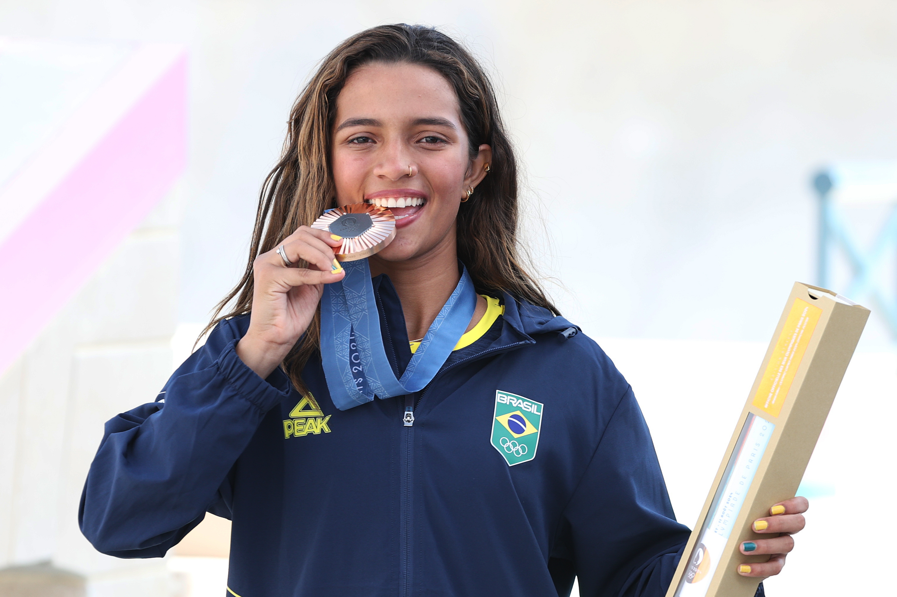 Atleta brasileira sorri e morde medalha de bronze dos Jogos Olímpicos de Paris 2024, vestindo uniforme do Brasil.
