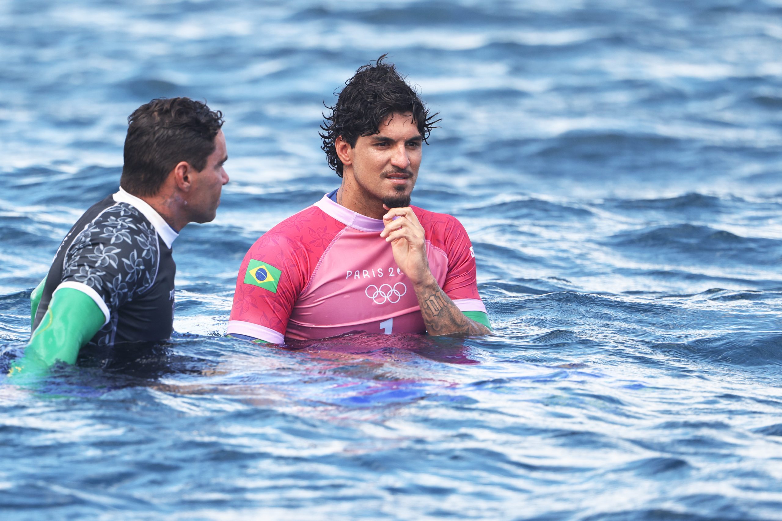 Dois atletas de surf na água, um com camisa rosa com bandeira do Brasil e logo de Paris 2024.