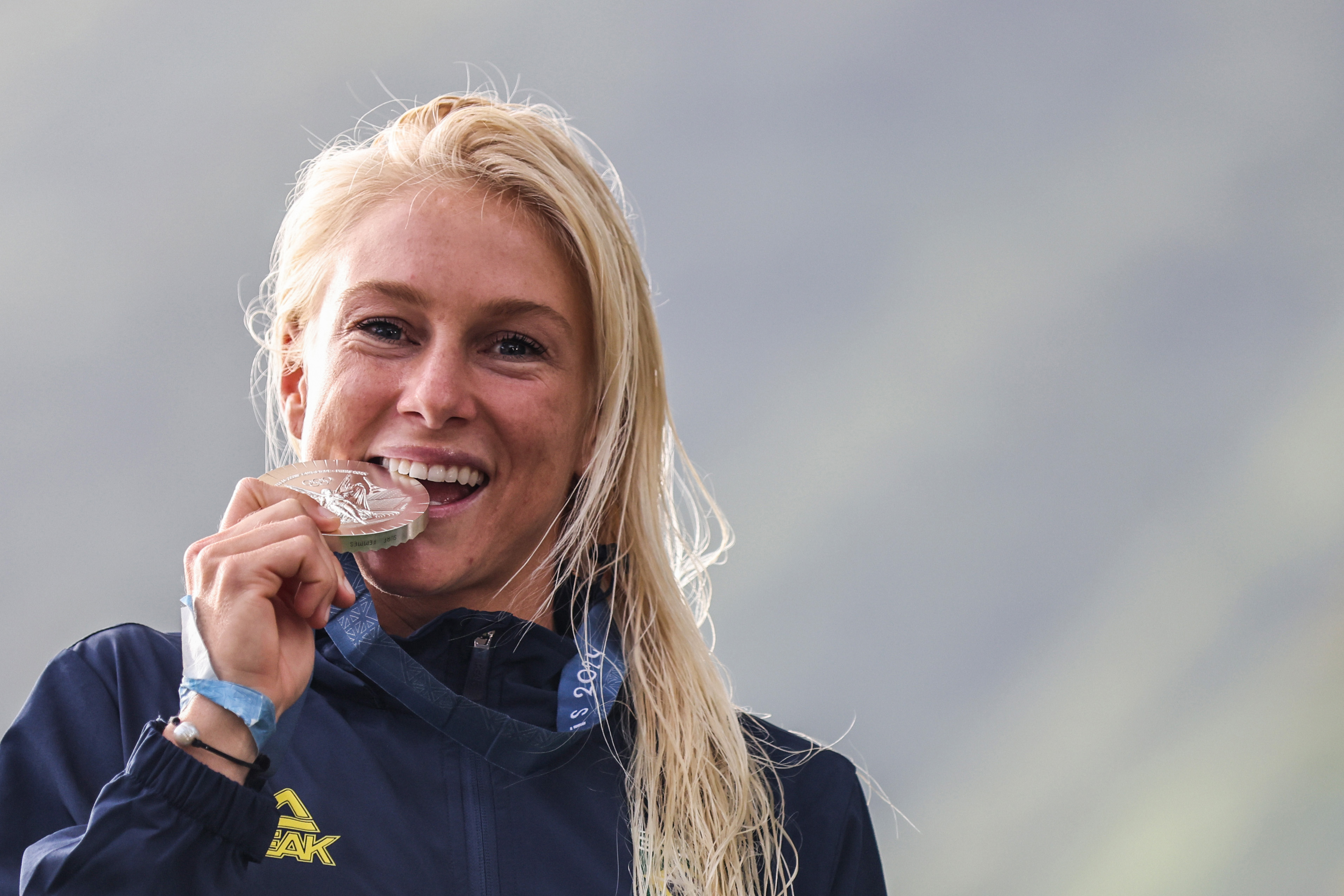 Atleta loira sorrindo, com cabelo molhado, mordendo uma medalha de prata.