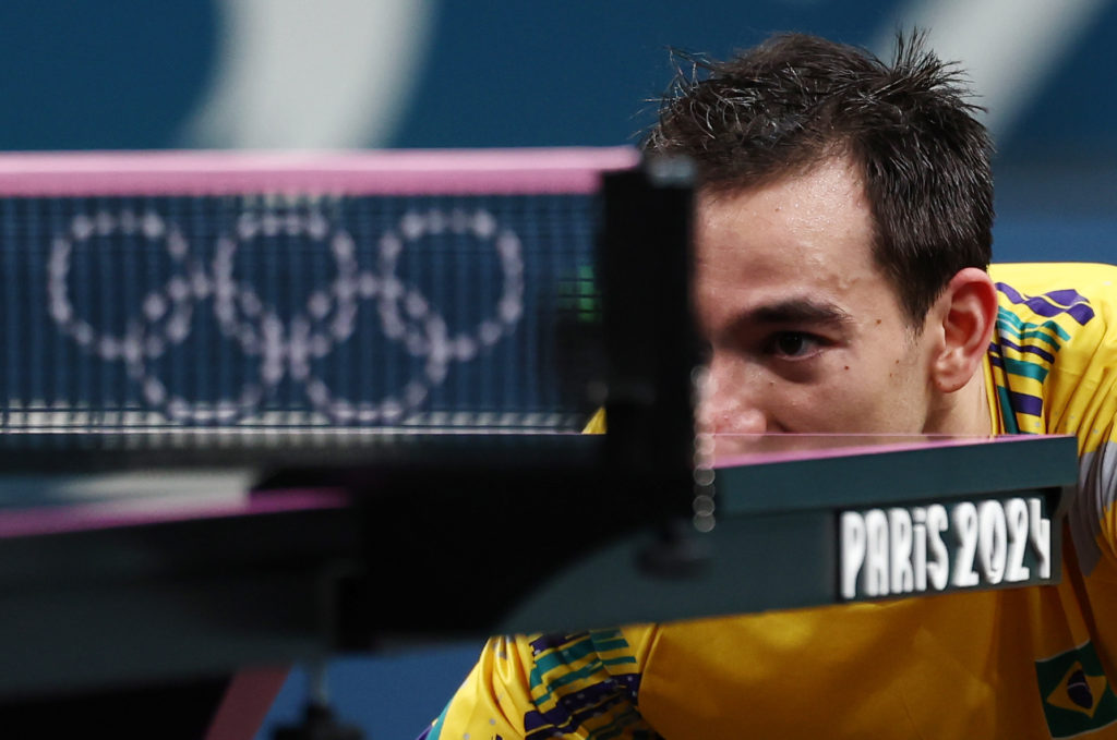Atleta de tênis de mesa brasileiro focado, olhando a rede. Anéis olímpicos e 'Paris 2024' em destaque.