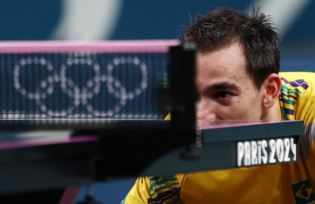 Atleta de tênis de mesa brasileiro focado, olhando a rede. Anéis olímpicos e 'Paris 2024' em destaque.