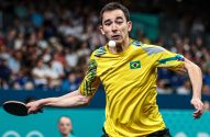 Jogador brasileiro de tênis de mesa se prepara para o saque, com a bola no ar perto do rosto. Camisa amarela.