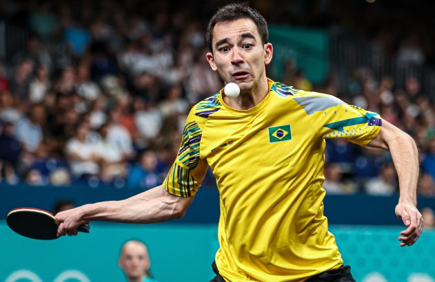 Jogador brasileiro de tênis de mesa se prepara para o saque, com a bola no ar perto do rosto. Camisa amarela.