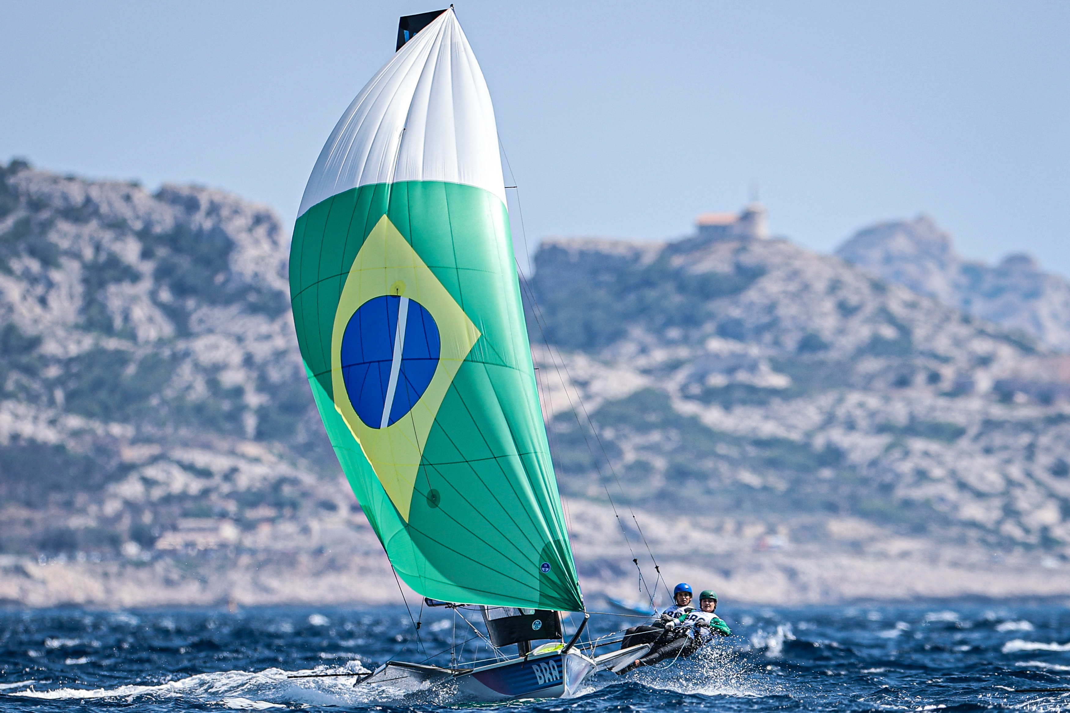 Veleiro de competição com vela da bandeira do Brasil, dois velejadores, navegando rápido em mar agitado.
