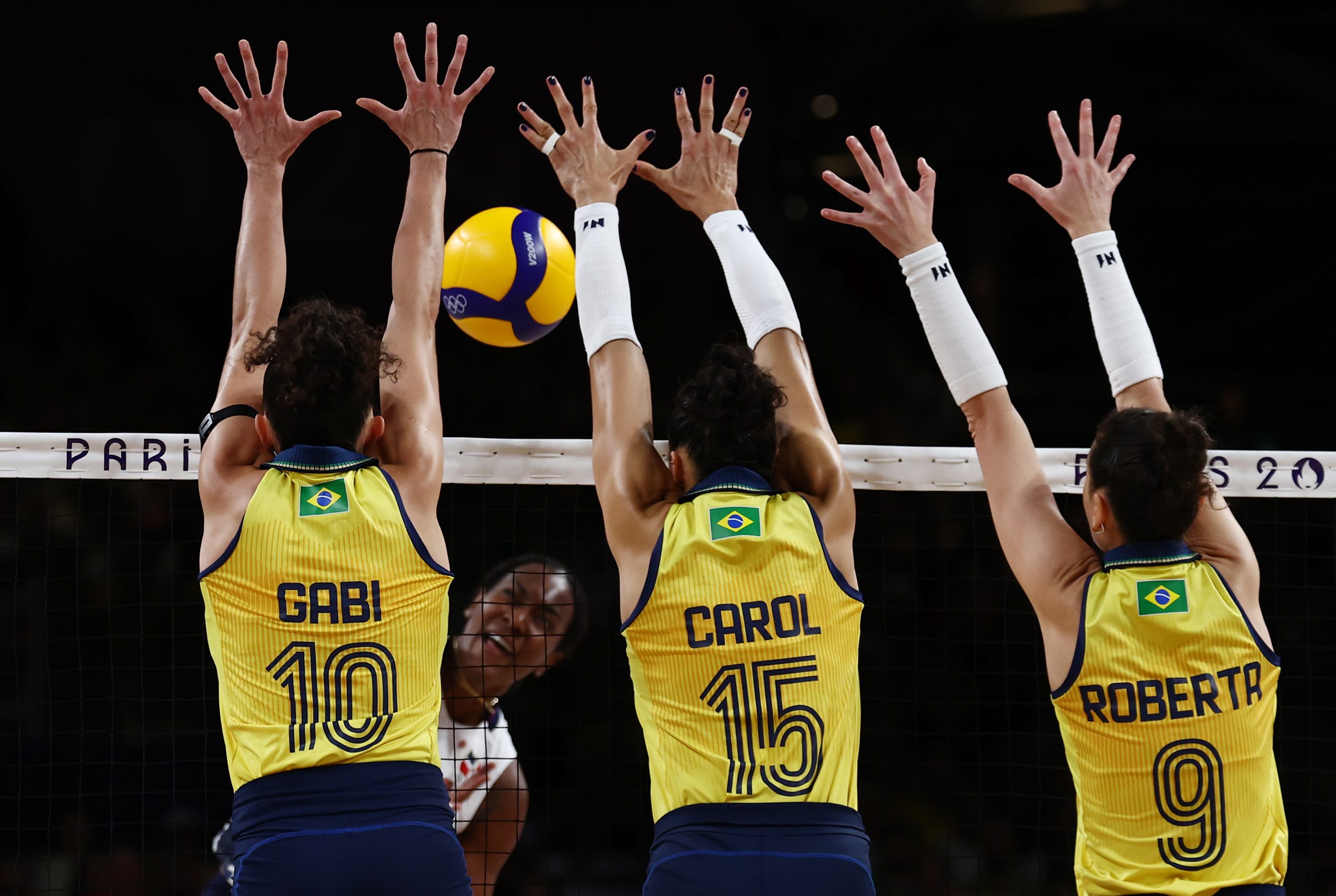 Três jogadoras de vôlei do Brasil (Gabi, Carol, Roberta) bloqueiam a bola na rede durante jogo. Fundo escuro, 'Paris 2024'.