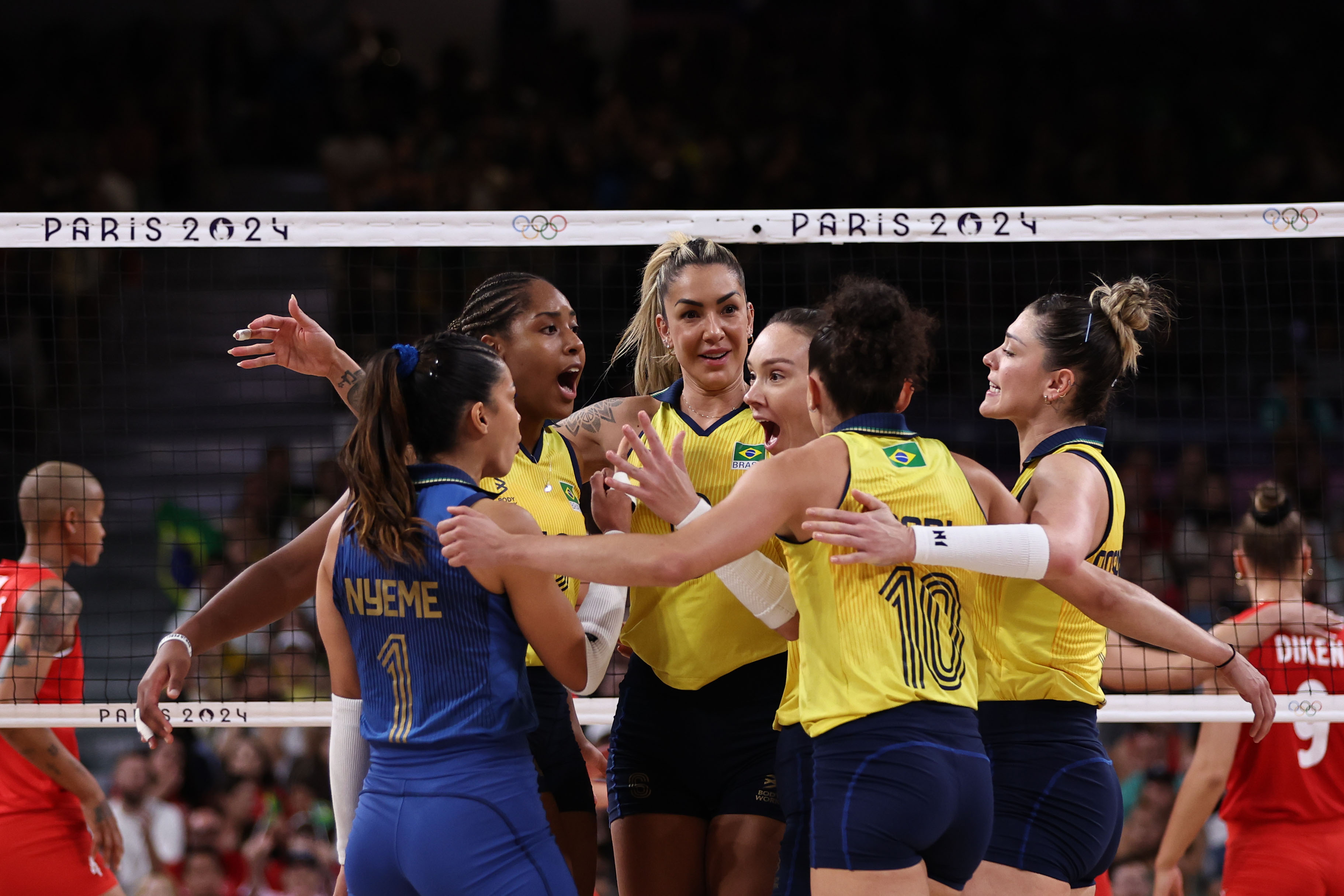 Vôlei: Jogadoras brasileiras celebram ponto em jogo. Usam uniformes amarelo/azul, com rede "PARIS 2024" ao fundo.