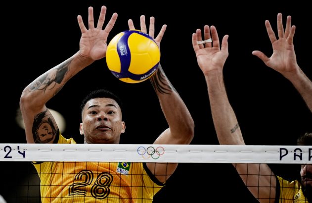 Jogador de vôlei de camisa amarela nº 28 bloqueia a bola sobre a rede, com outros dois bloqueadores ao lado.