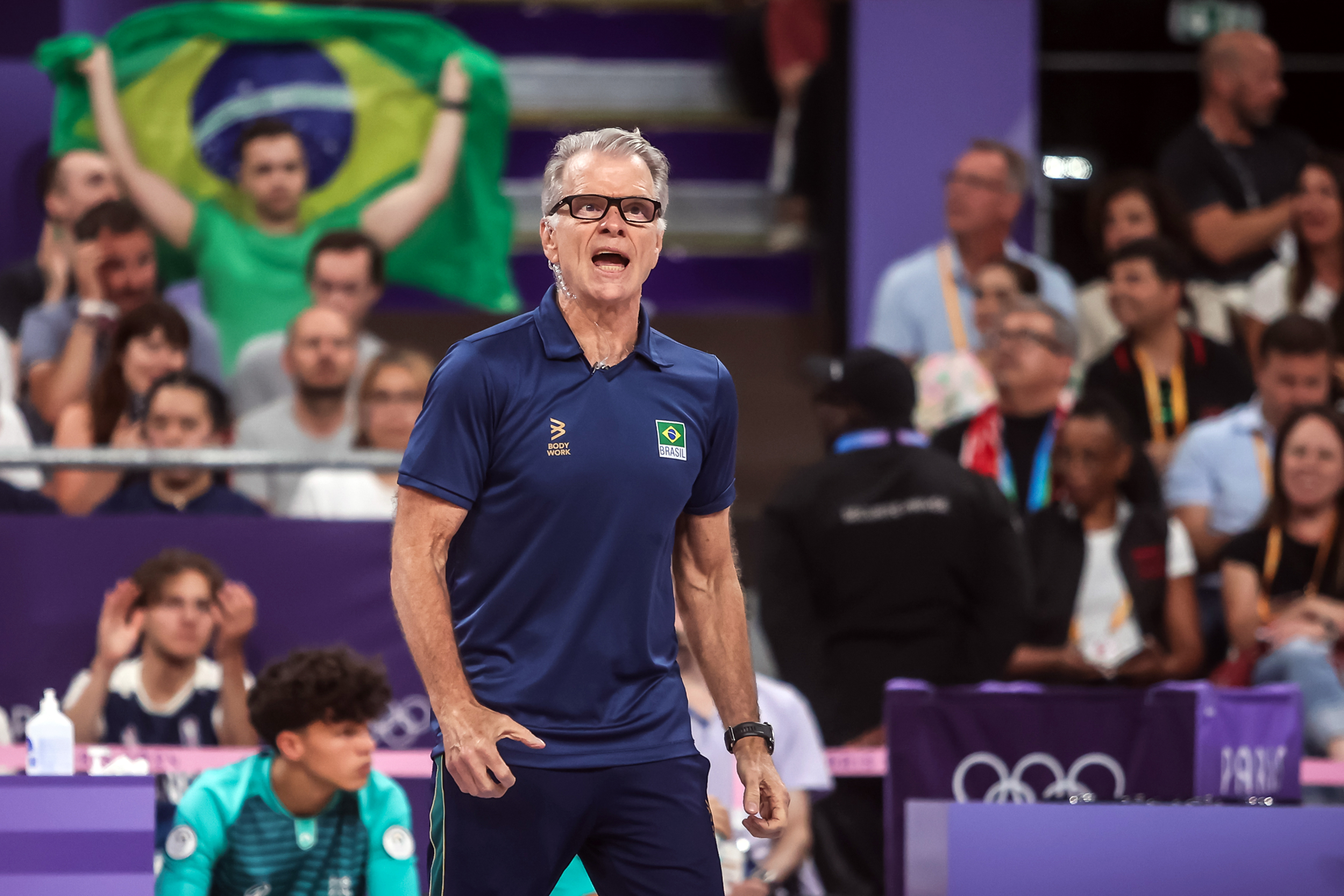 Homem em camisa da seleção brasileira grita, com torcida exibindo a bandeira do Brasil ao fundo.