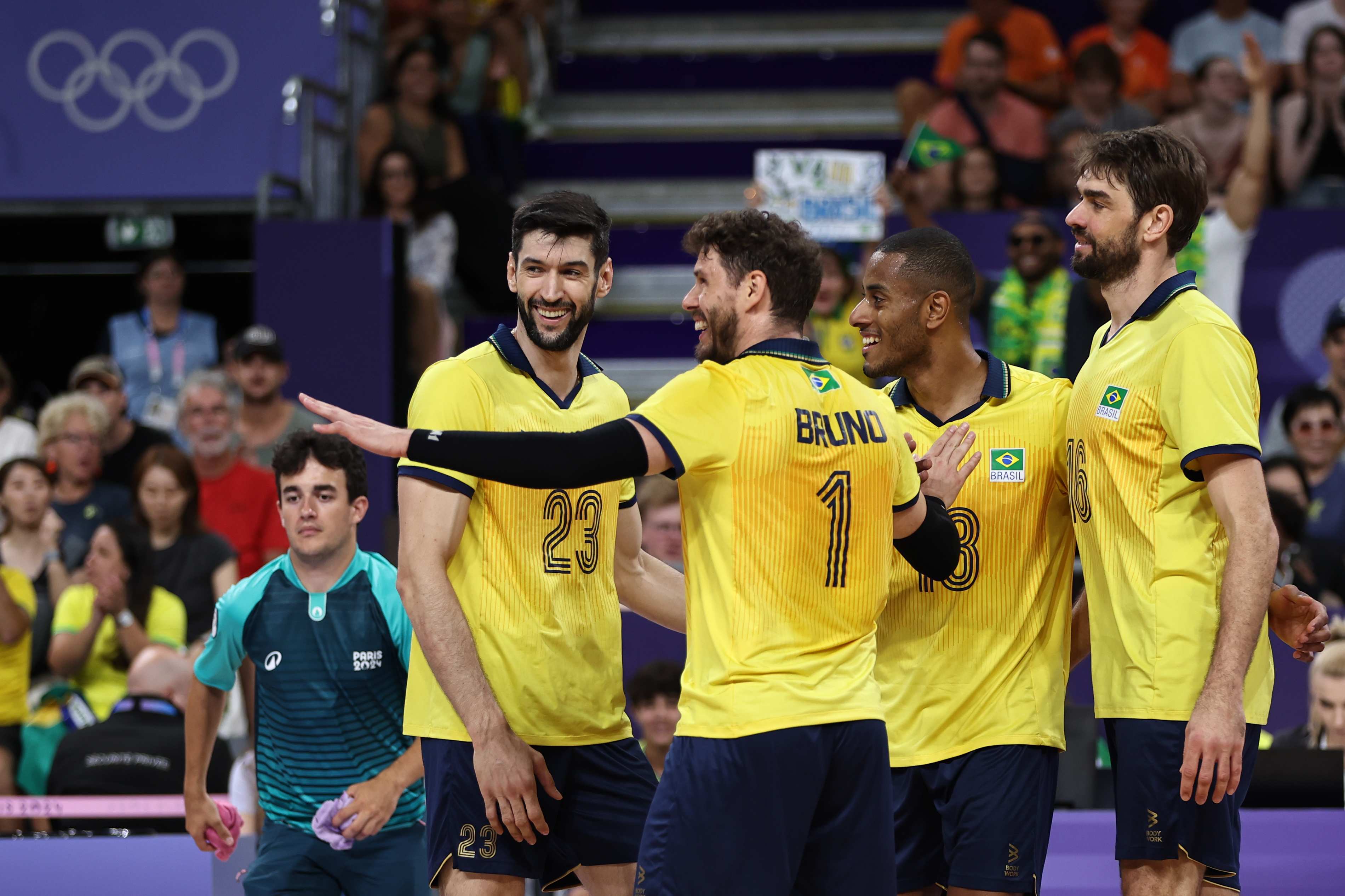 Jogadores de vôlei do Brasil em uniformes amarelos se reúnem sorrindo na quadra, com anéis olímpicos ao fundo.