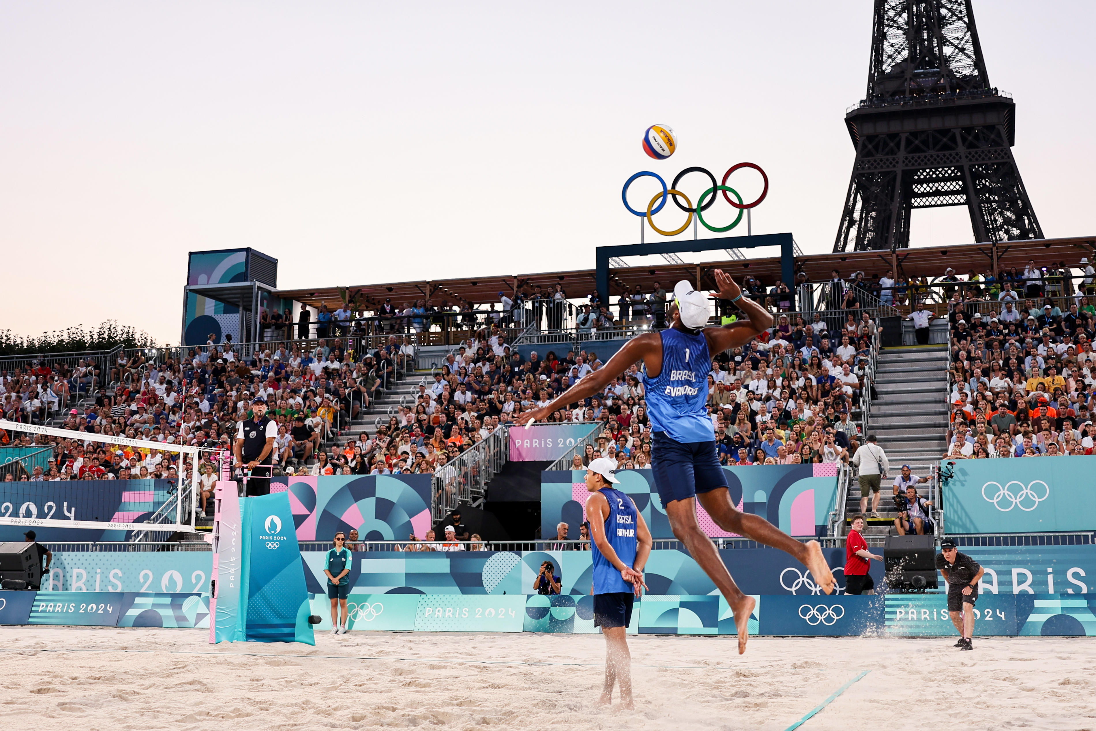 Jogador de vôlei de praia do Brasil salta para cortar a bola. Anéis Olímpicos e Torre Eiffel ao fundo.