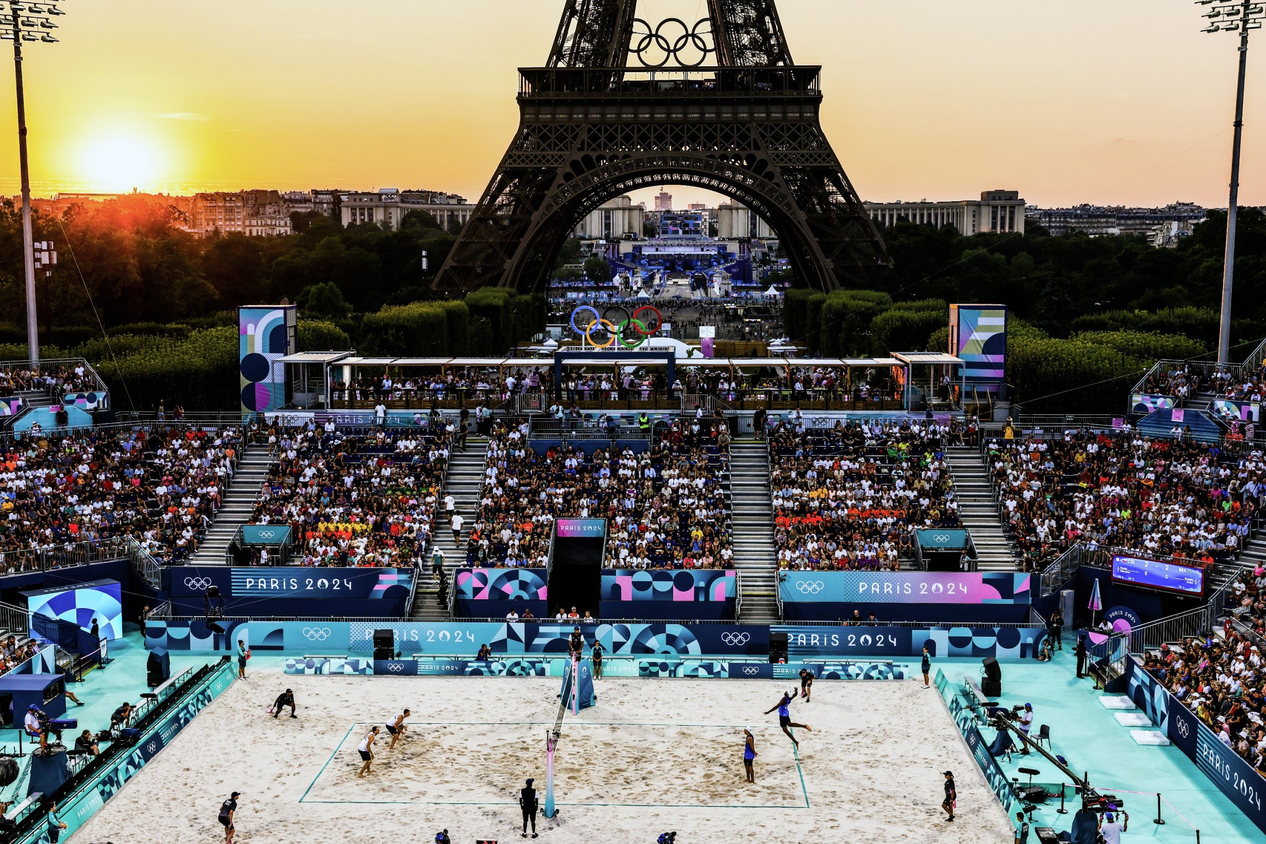 Partida de vôlei de praia dos Jogos Paris 2024 ao pôr do sol, com a Torre Eiffel, anéis olímpicos e torcedores.