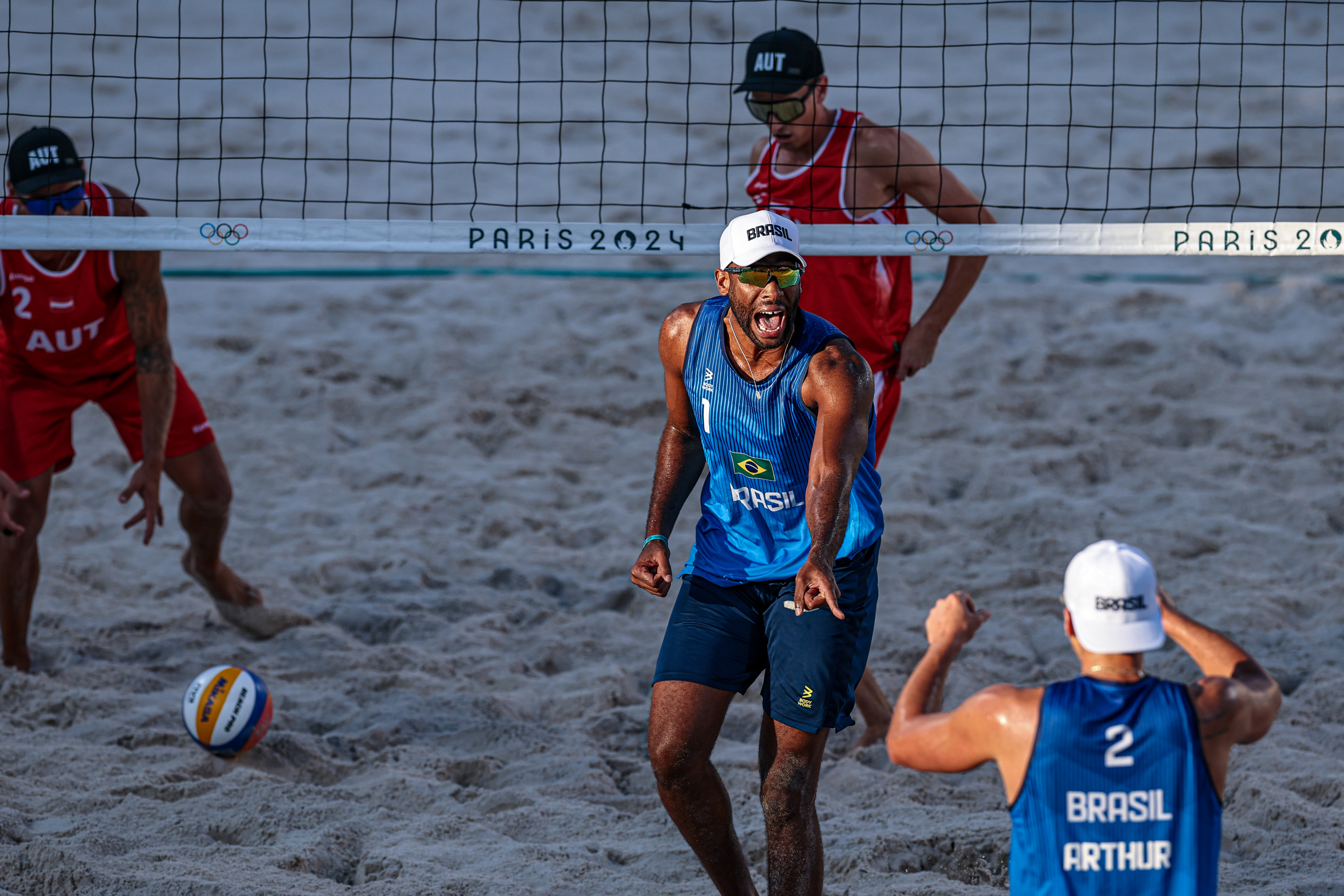 Vôlei de praia. Jogadores do Brasil celebram um ponto, enquanto equipe da Áustria se posiciona. Rede com "PARIS 2024".