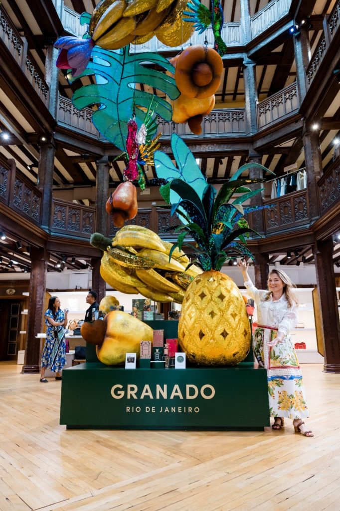 Mulher posa em display da Granado com frutas gigantes em espaço de madeira ornamentado.