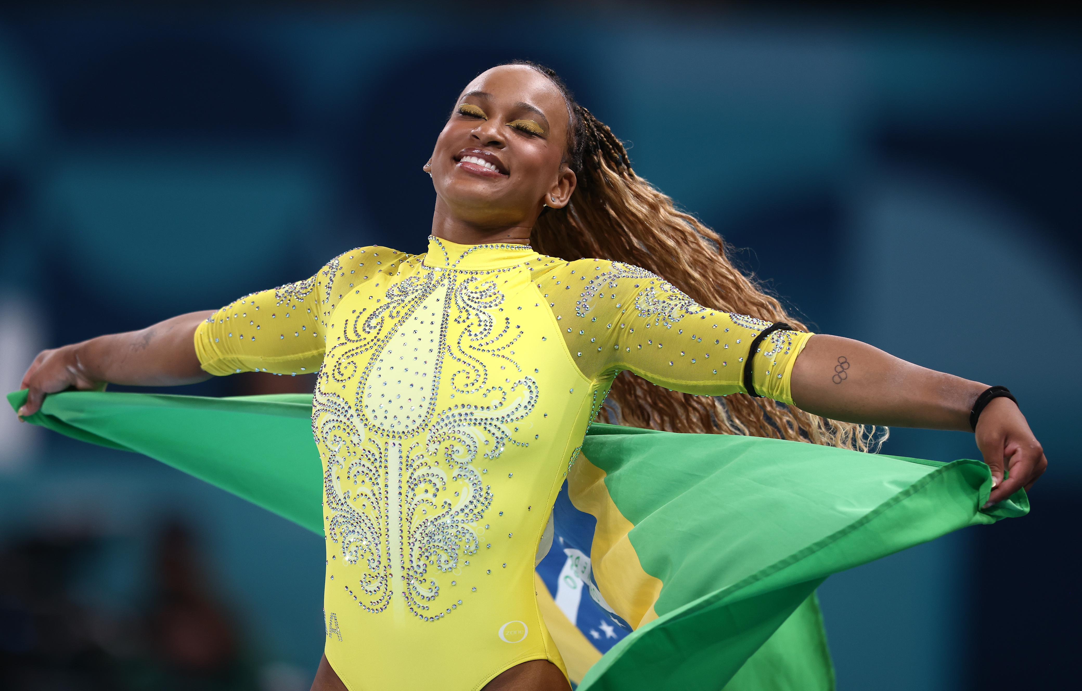 Ginasta sorridente em leotard amarelo brilhante, segurando a bandeira do Brasil com os braços abertos.