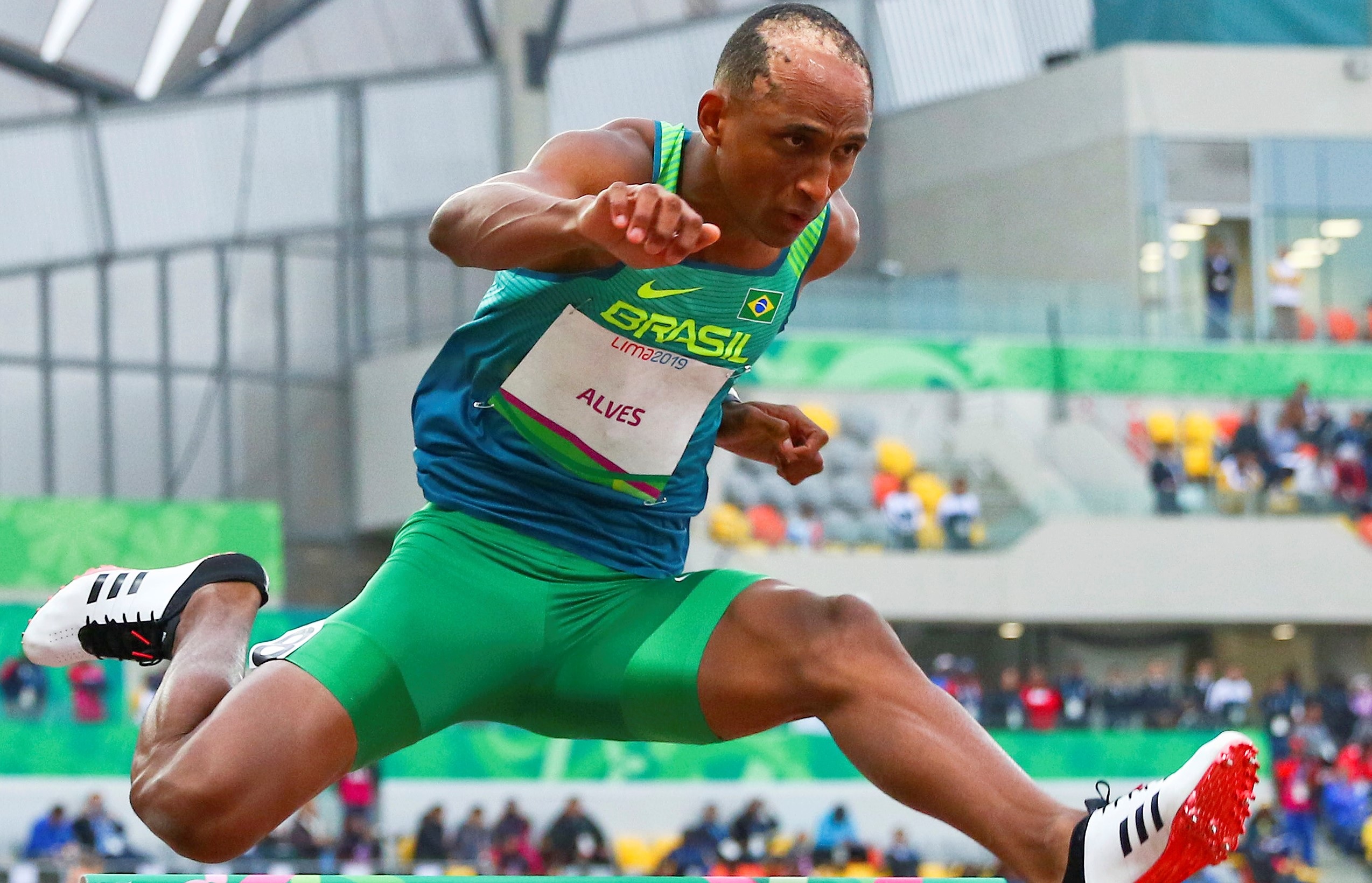 Atleta brasileiro Alves salta barreira, vestindo uniforme verde e azul do Brasil, Lima 2019.