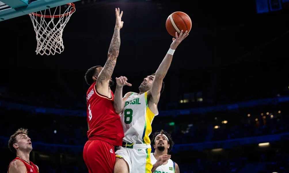 Basquete: jogador de camisa branca/verde faz bandeja. Defensor de vermelho tenta bloquear perto da cesta.