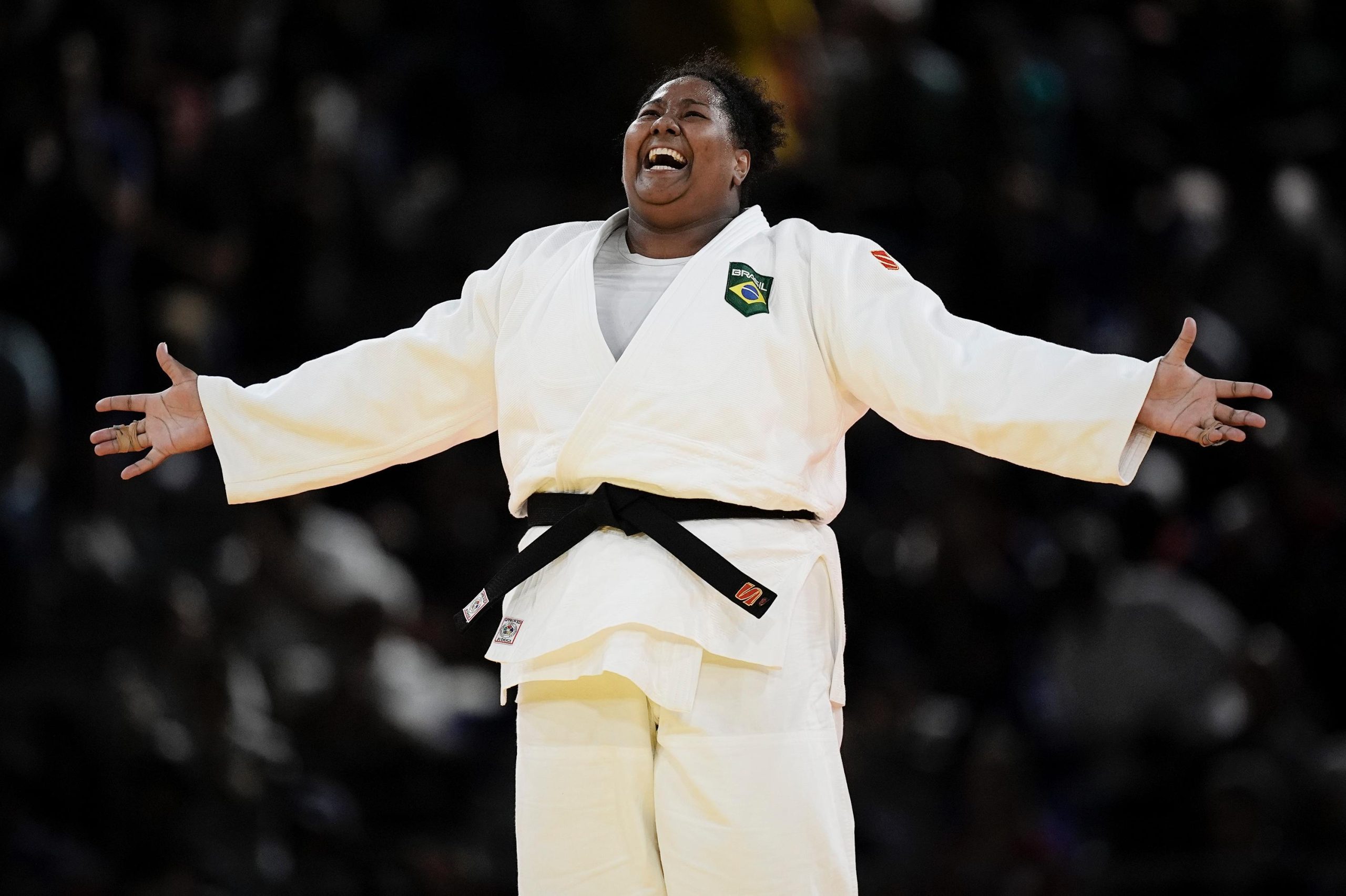 Judoca brasileira com judogi branco e faixa preta celebra com braços abertos e rosto de triunfo.