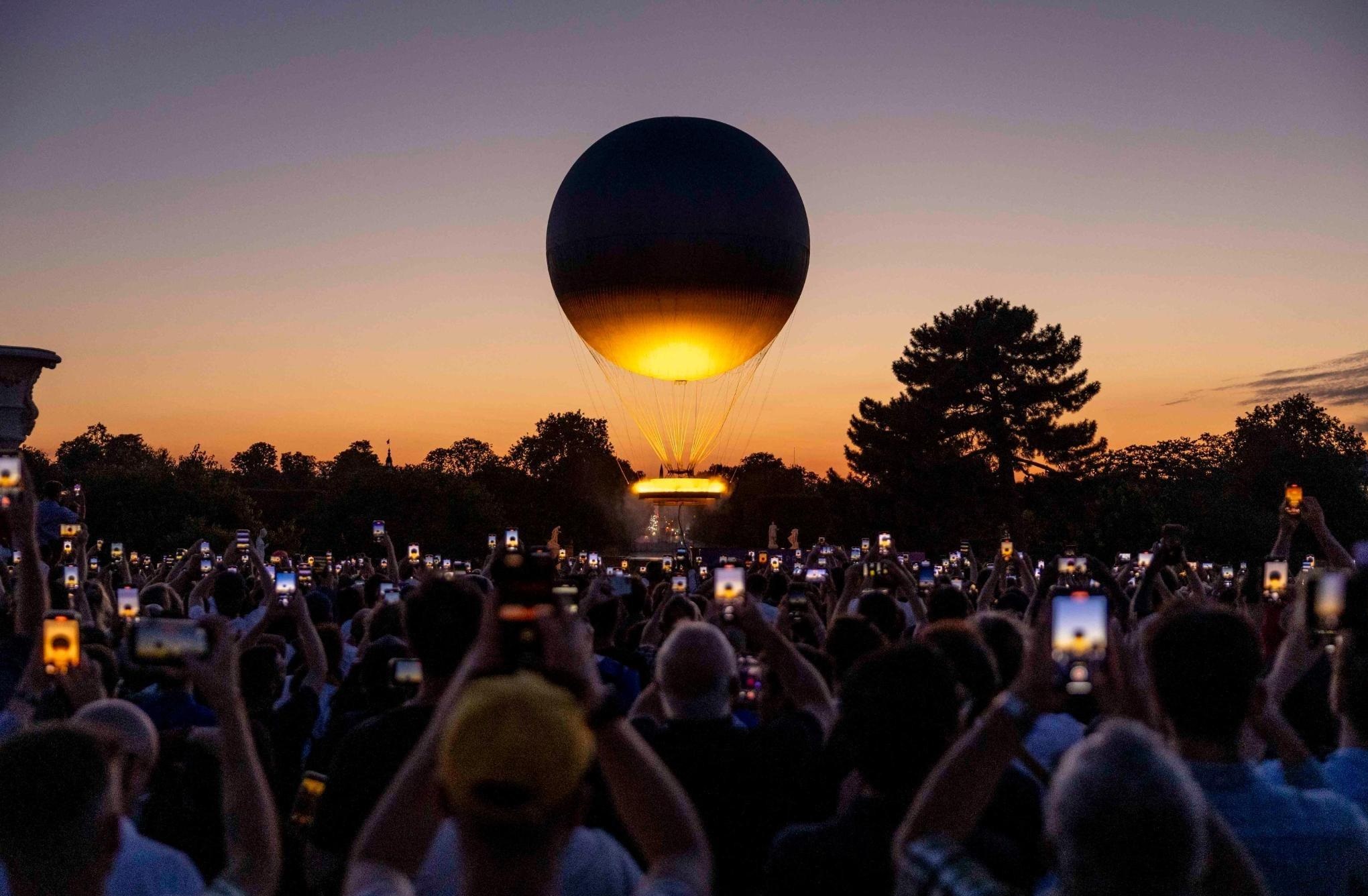 Balão esférico iluminado no crepúsculo. Multidão filma com celulares erguidos.