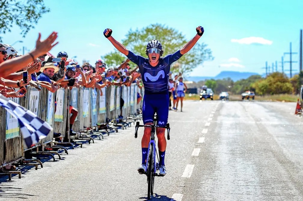 Ciclista em uniforme azul celebra vitória, braços erguidos, na linha de chegada. Multidão e bandeira quadriculada.