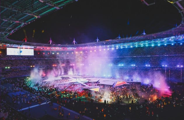 Estádio à noite, com público, luzes coloridas e confetes sendo lançados no palco de um evento vibrante.